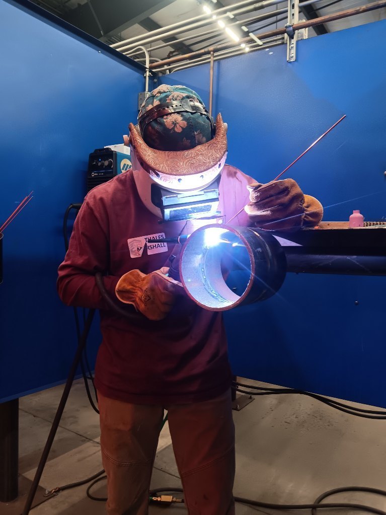 Students participating in a welding competition, wearing protective welding helmets and gear while working on pipe welding projects in a shop environment. 3