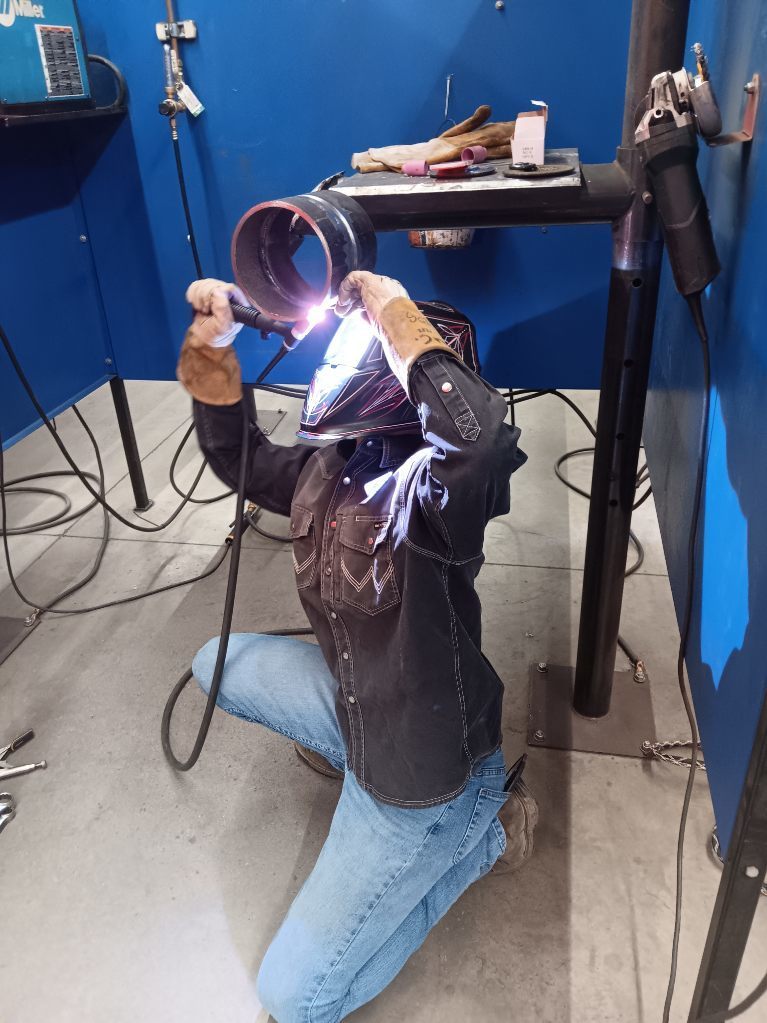 Students participating in a welding competition, wearing protective welding helmets and gear while working on pipe welding projects in a shop environment. 2