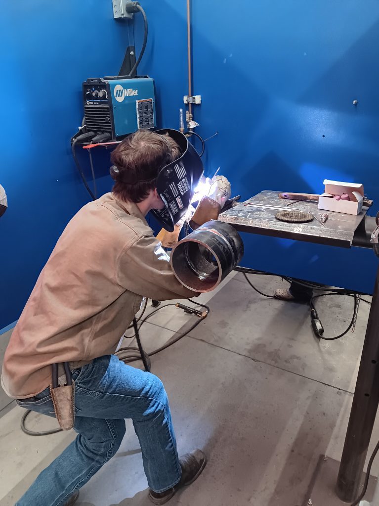 Students participating in a welding competition, wearing protective welding helmets and gear while working on pipe welding projects in a shop environment. 4