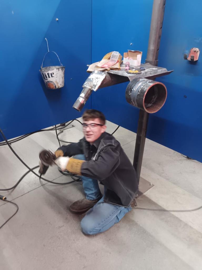 Students participating in a welding competition, wearing protective welding helmets and gear while working on pipe welding projects in a shop environment. 5