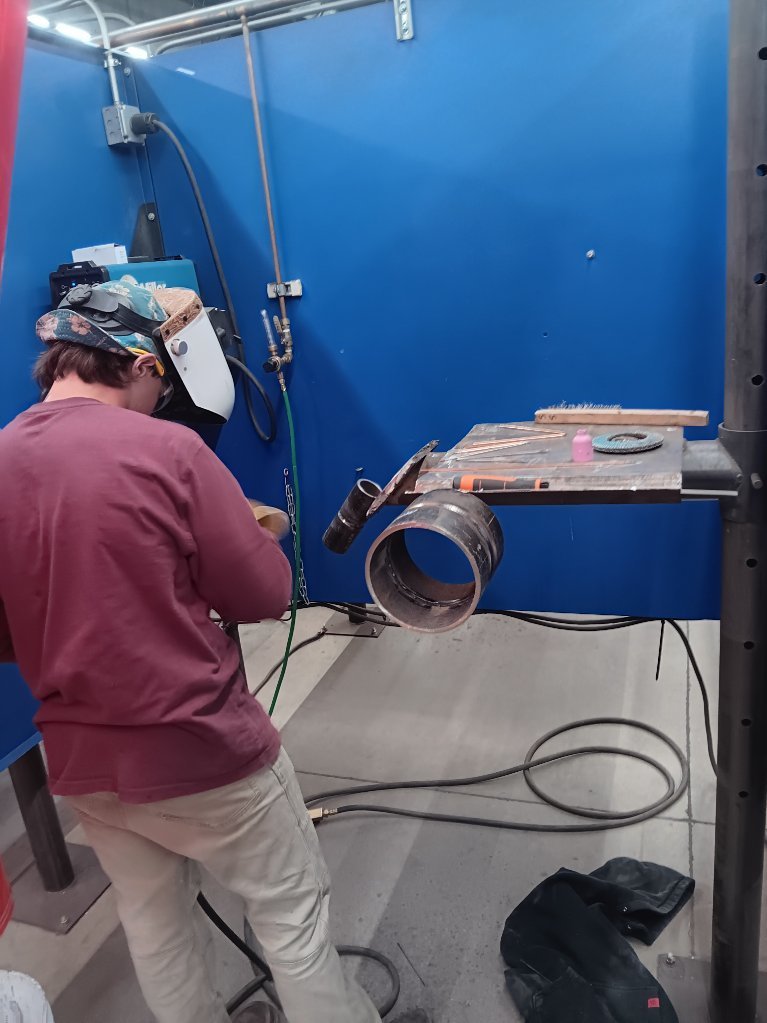 Students participating in a welding competition, wearing protective welding helmets and gear while working on pipe welding projects in a shop environment. 6
