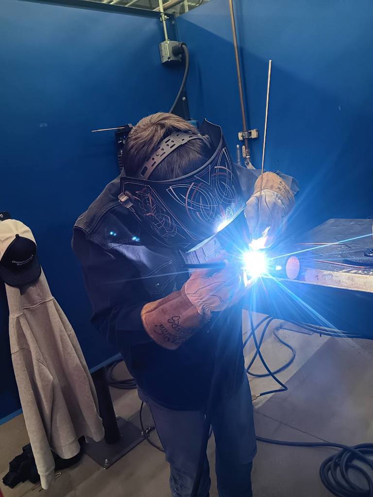 Students participating in a welding competition, wearing protective welding helmets and gear while working on pipe welding projects in a shop environment. 7