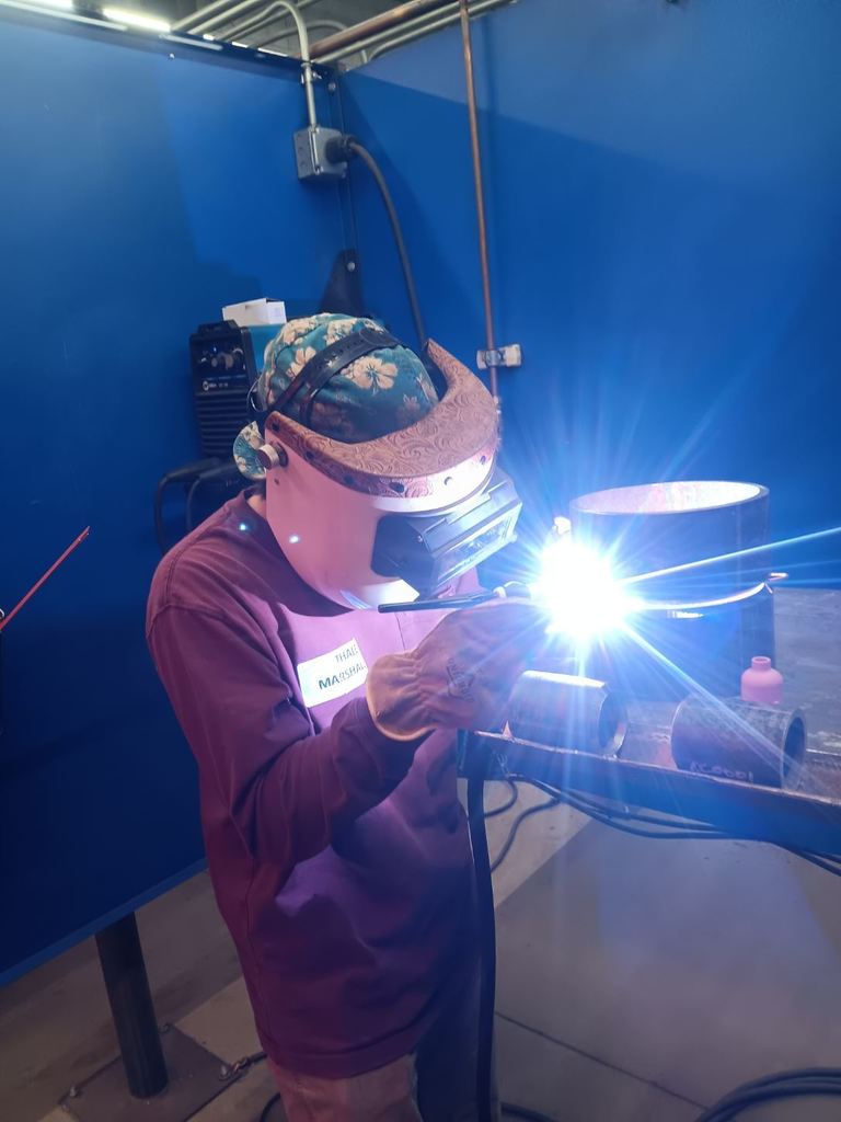 Students participating in a welding competition, wearing protective welding helmets and gear while working on pipe welding projects in a shop environment. 8
