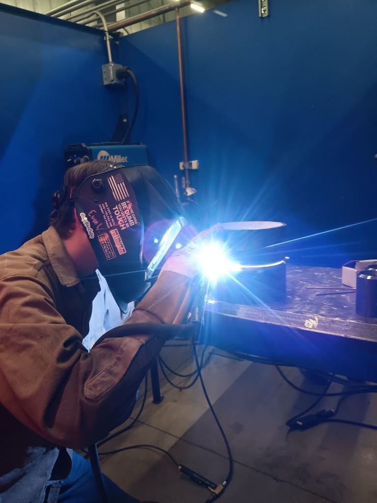 Students participating in a welding competition, wearing protective welding helmets and gear while working on pipe welding projects in a shop environment. 9