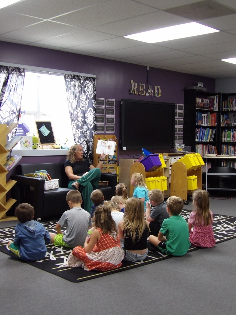 Elementary students getting read a book in the library by elementary teacher.