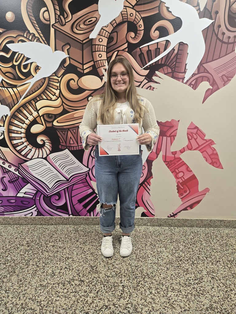 Katelyn is holding up a student of the month sign while wearing a cream colored sweater, blue jeans and white sneakers. 