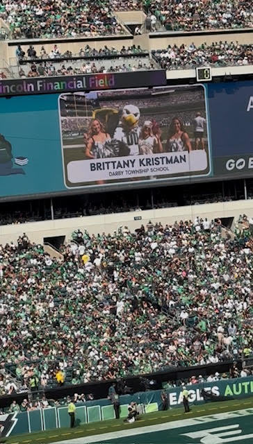 A stadium jumbotron shows a photo of Brittany Kristman standing with two others and the Eagles mascot. The text on the screen reads Brittany Kristman, Darby Township School. The crowd below fills the stands in green jerseys.