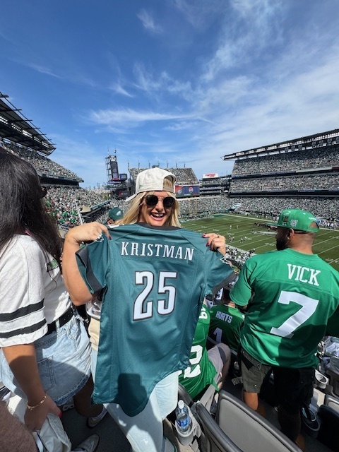 A smiling woman at a football stadium holds up a green Eagles jersey with the name Kristman and the number 25. The stands are filled with fans wearing green, and the field is visible in the background.