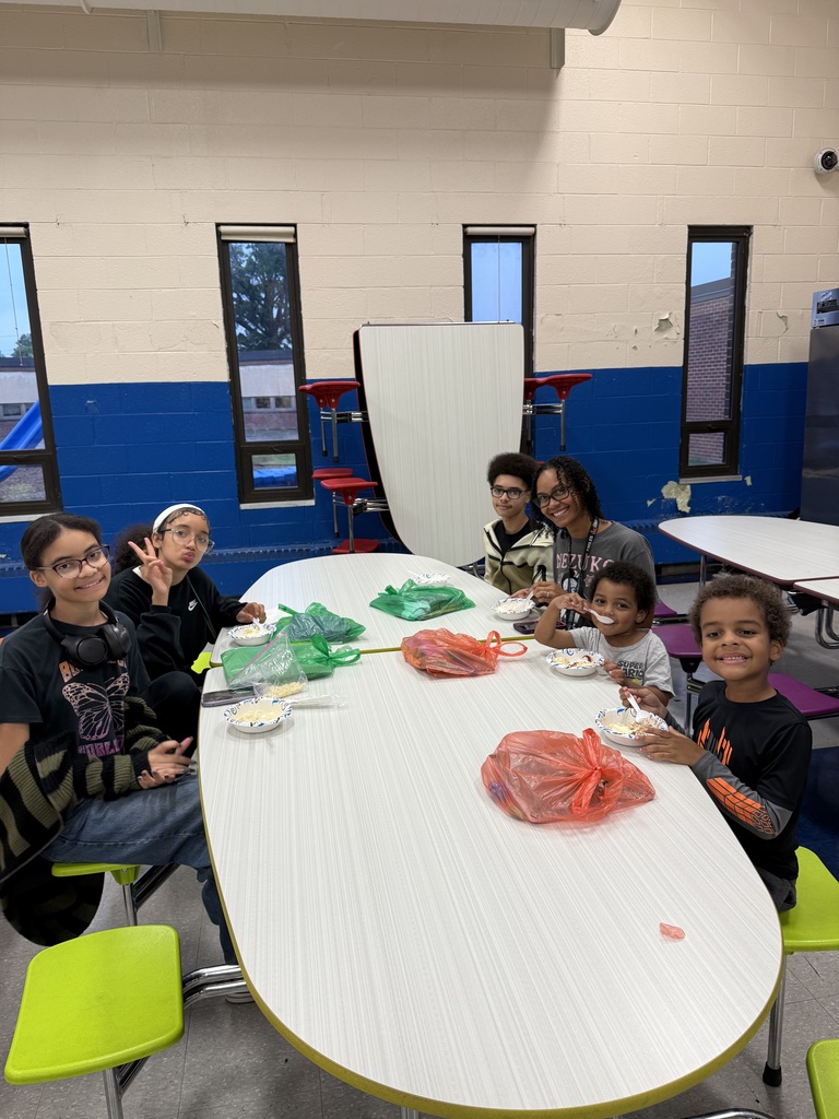 A group of children and an adult sit at a cafeteria table, smiling and eating.