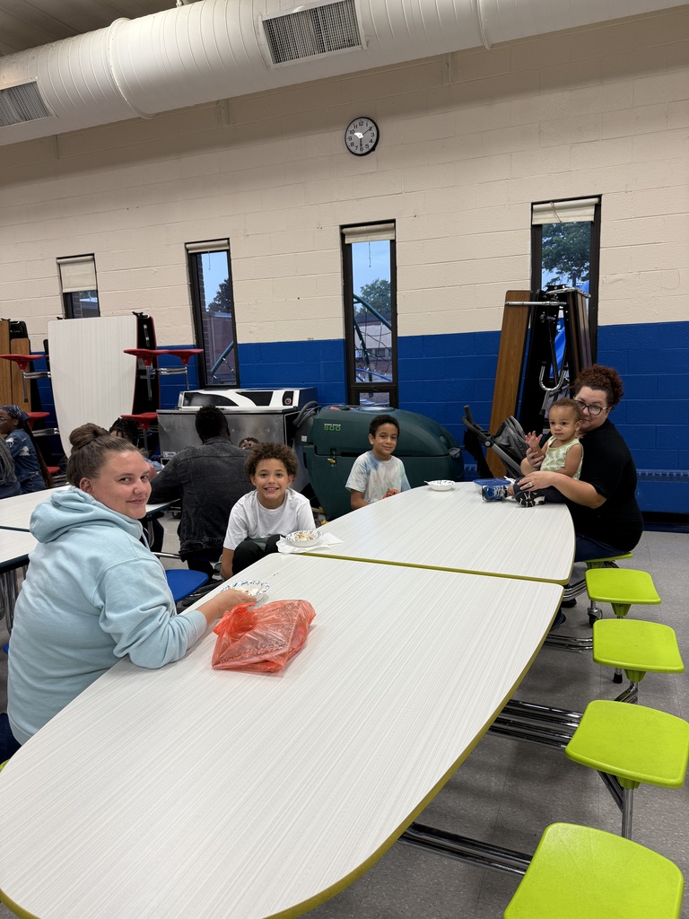 A woman in a light blue sweatshirt smiles at the camera, with children and another adult seated nearby.