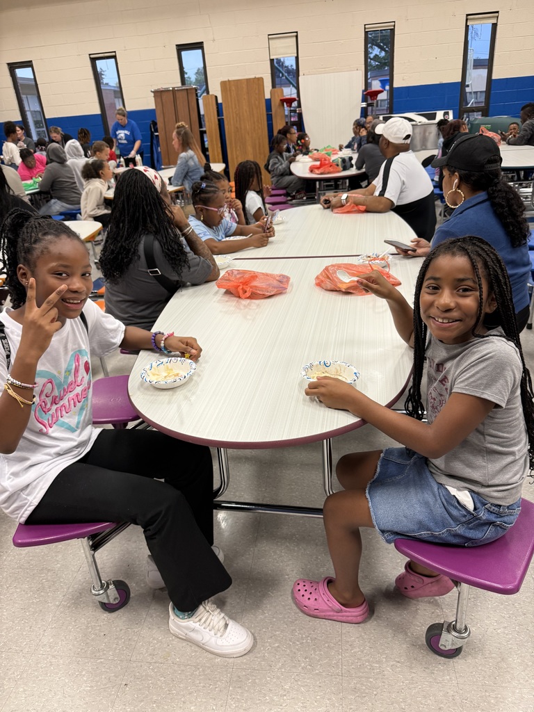 Two young girls smile at the camera while eating at a cafeteria table. One holds up a peace sign and the other smiles with a spoon.