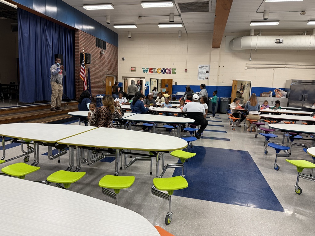 Families sit at cafeteria tables while a man speaks on stage in front of a blue curtain and U.S. flag.