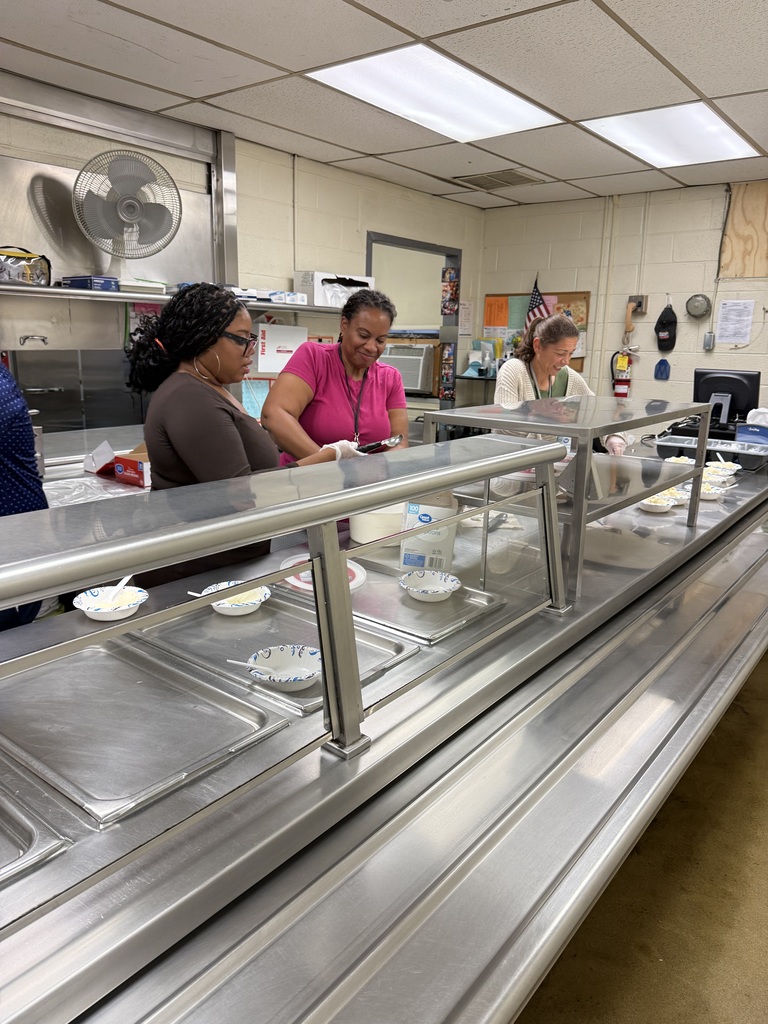 Three women stand behind a cafeteria serving line, preparing and serving ice cream into bowls.