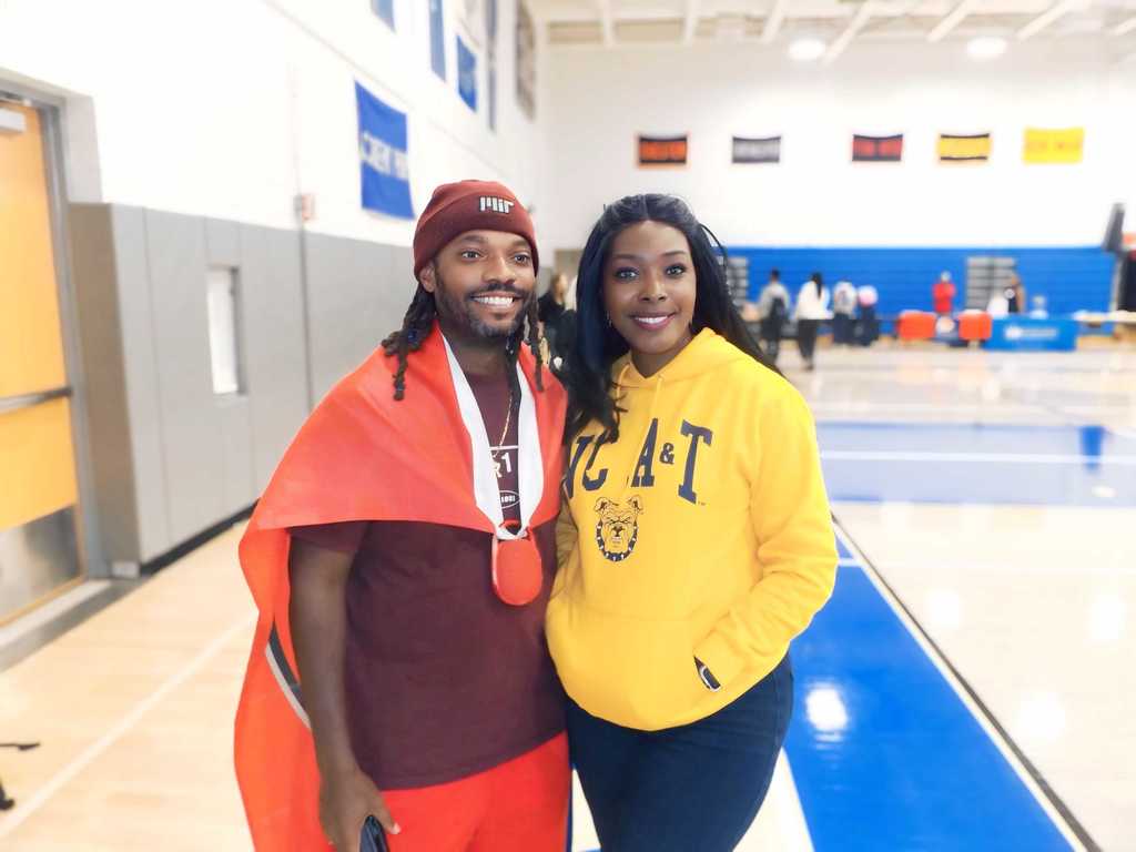 Two adults smile for the camera in the gym. One wears a red Harvard shirt and beanie, the other wears a yellow NC A&T hoodie.