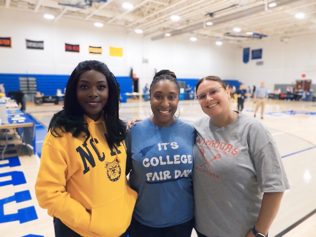 Three women smile for a photo in the gym. One wears an NC A&T hoodie, one wears a shirt that says 'It’s College Fair Day,' and one wears a Warriors lacrosse shirt.