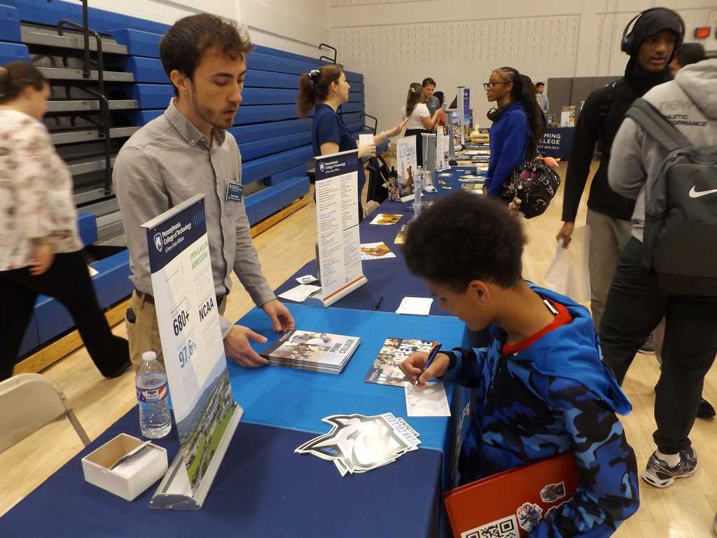 A student fills out a form at a college fair table while a representative explains admissions information.