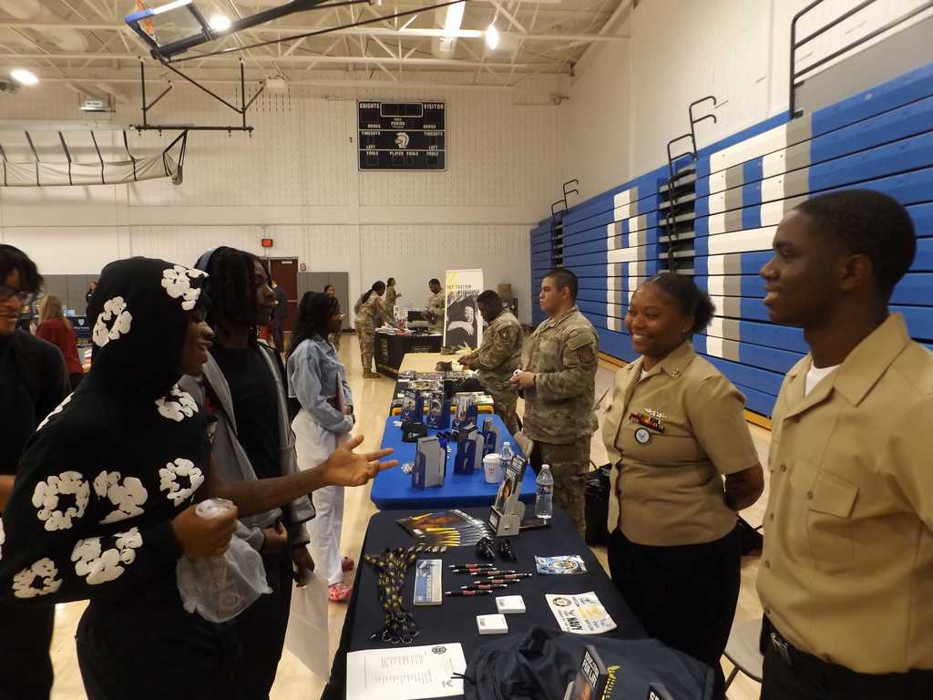 Students speak with ROTC representatives at a table during a college fair held in the school gym.