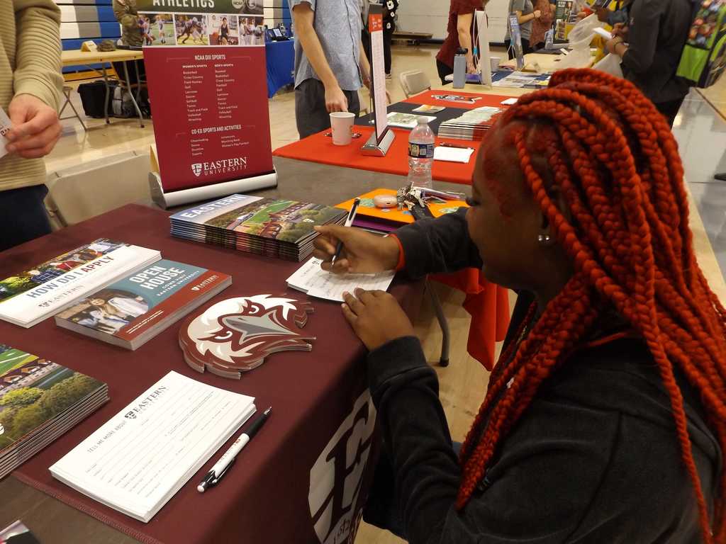 A student with red braids writes on a form at the Eastern University table, which displays brochures, pamphlets, and athletics information.