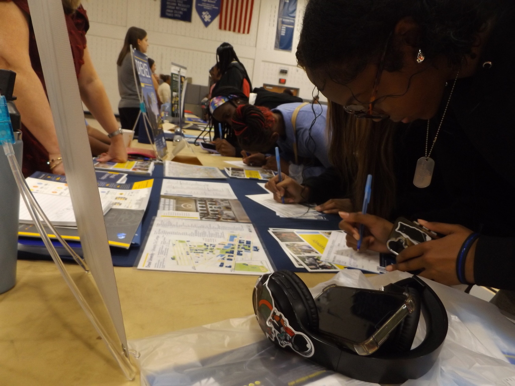 Students lean over a table, writing on forms and gathering information during a college fair.