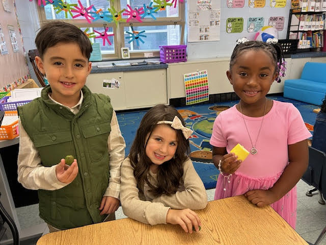 Three young students stand at a classroom table holding pieces of fruit and smiling