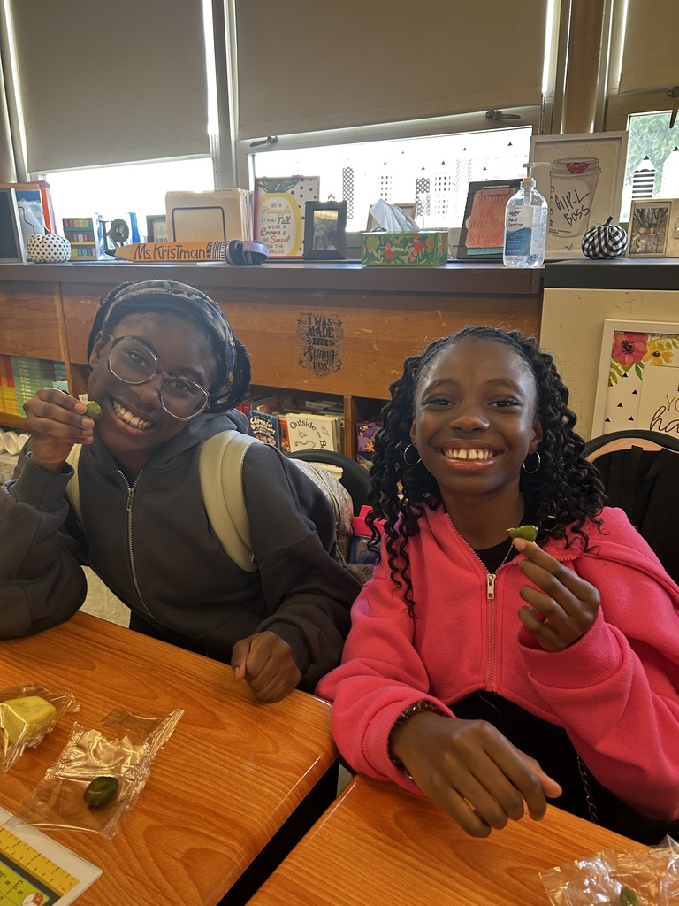 Two smiling students sit at a classroom desk holding pieces of fruit, posing happily for the camera