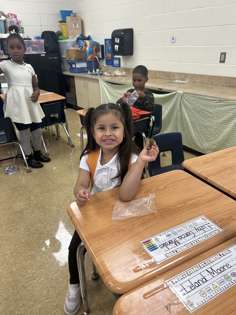 A young student sits at a desk holding up a piece of fruit and smiling