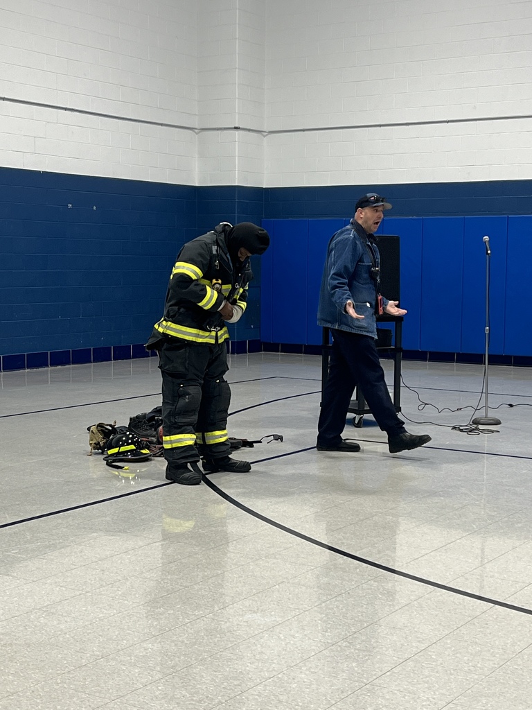 A firefighter in protective gear and another presenter stand at the front of the gym. The presenter speaks at a microphone while the firefighter adjusts equipment on their uniform.