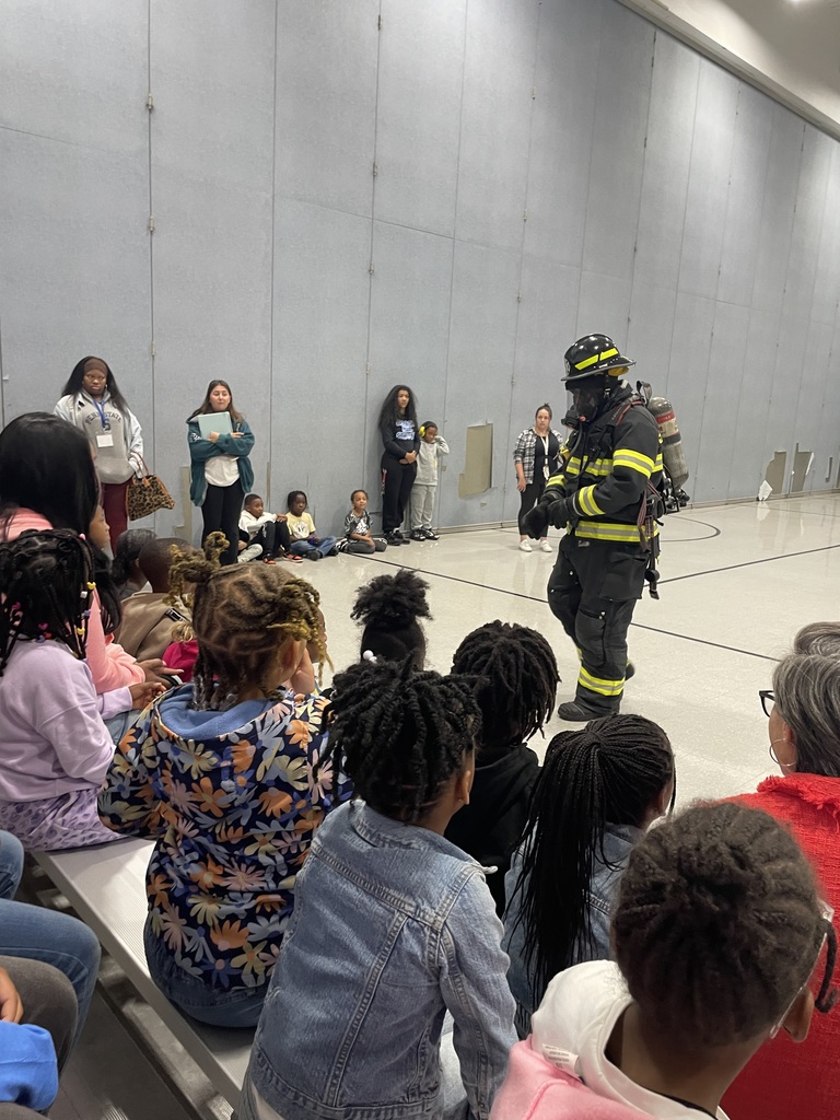 A firefighter in full gear, including helmet and oxygen tank, walks in front of seated elementary students during a school assembly.