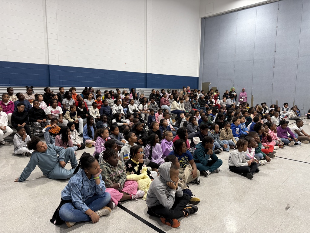 A large group of elementary students sit closely together on the floor of a gymnasium during an assembly. Teachers and staff are seated or standing along the back rows.