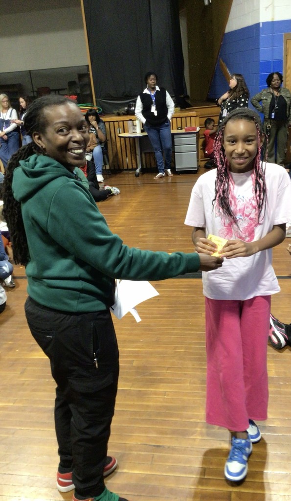 A smiling staff member in a green hoodie presents a certificate or award to a student wearing a pink outfit. They stand in a school gymnasium with other staff and students watching in the background.