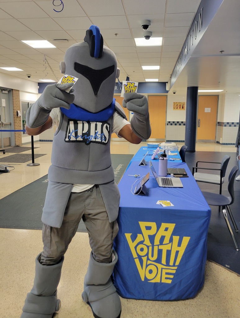 The school’s knight mascot poses in a hallway holding “PA Youth Vote” stickers, standing beside a blue table covered with matching “PA Youth Vote” materials.