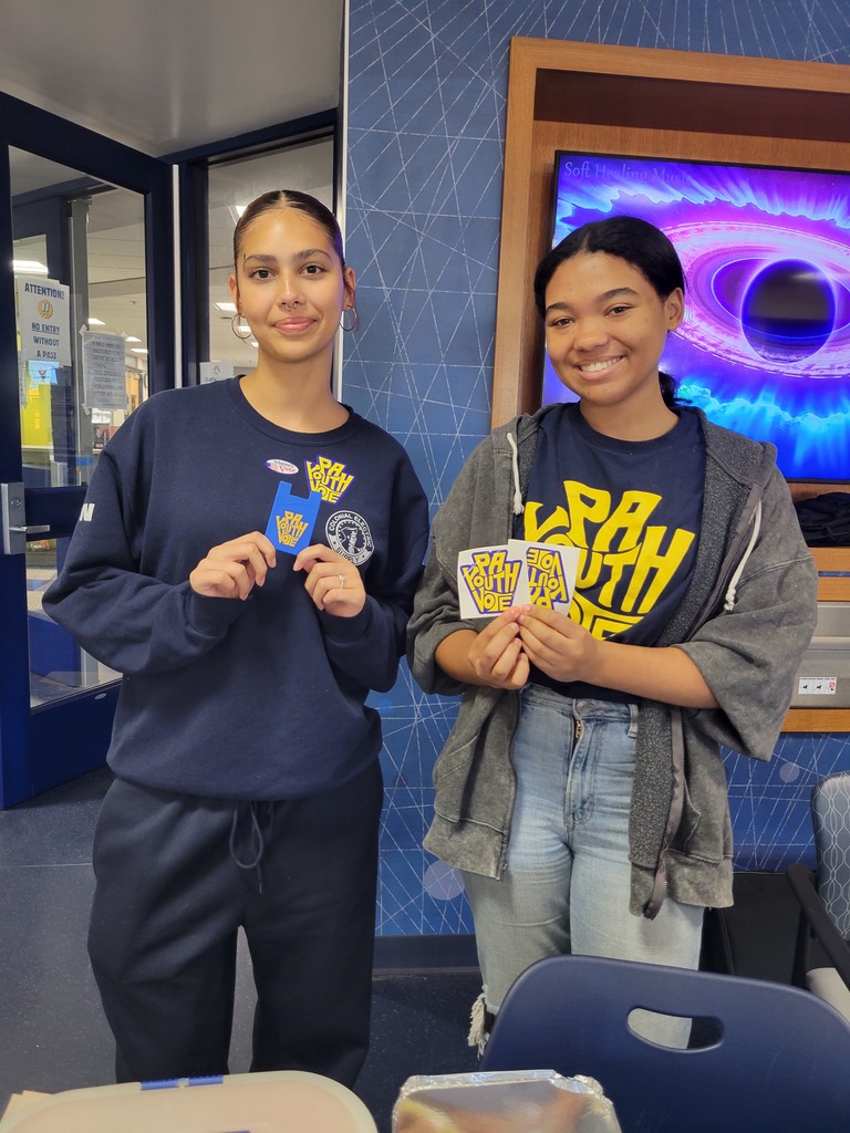 Two students stand behind a table, both smiling and holding several “PA Youth Vote” stickers.
