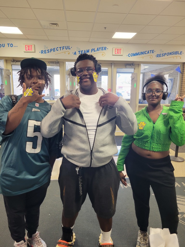 Three high school students stand in a hallway decorated with positive messages like “Be respectful” and “Be responsible.” They smile and show off “PA Youth Vote” stickers on their faces and hands.