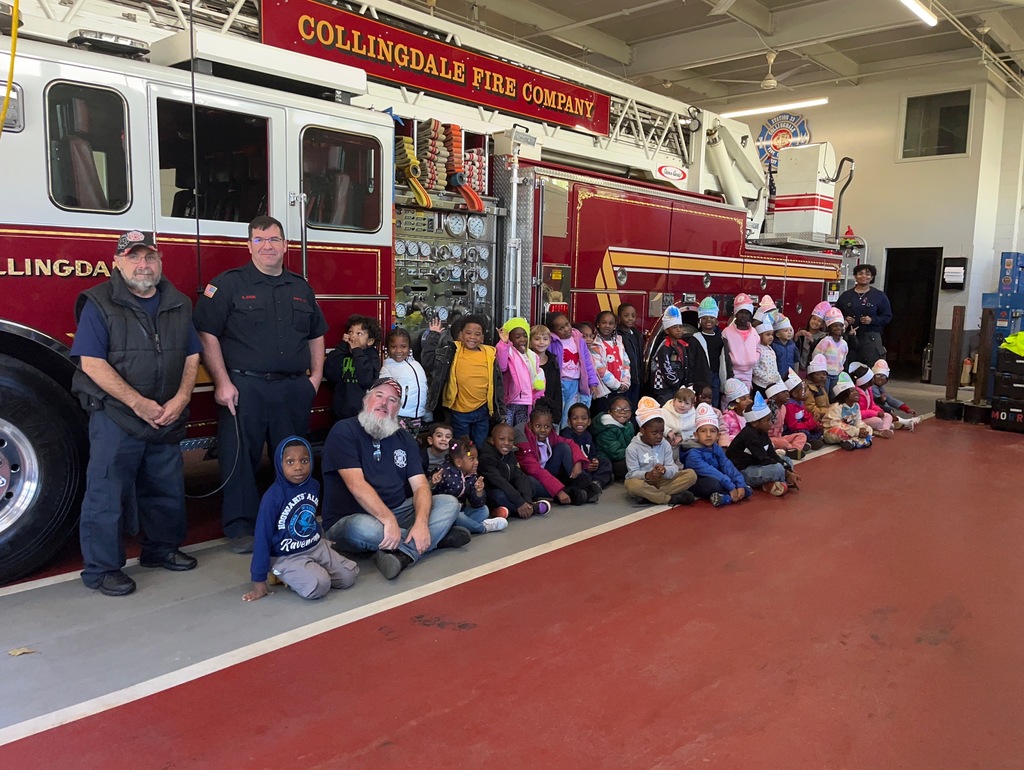 A large group of young students and several firefighters pose in front of a red Collingdale Fire Company fire truck inside the station. The children are seated or standing in rows, smiling and wearing paper firefighter hats.