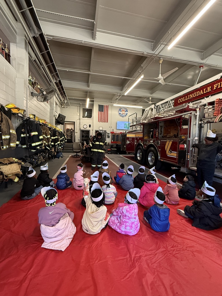 A group of young students wearing paper firefighter hats sit on a large red mat facing firefighters who are demonstrating equipment inside the Collingdale Fire Company station.