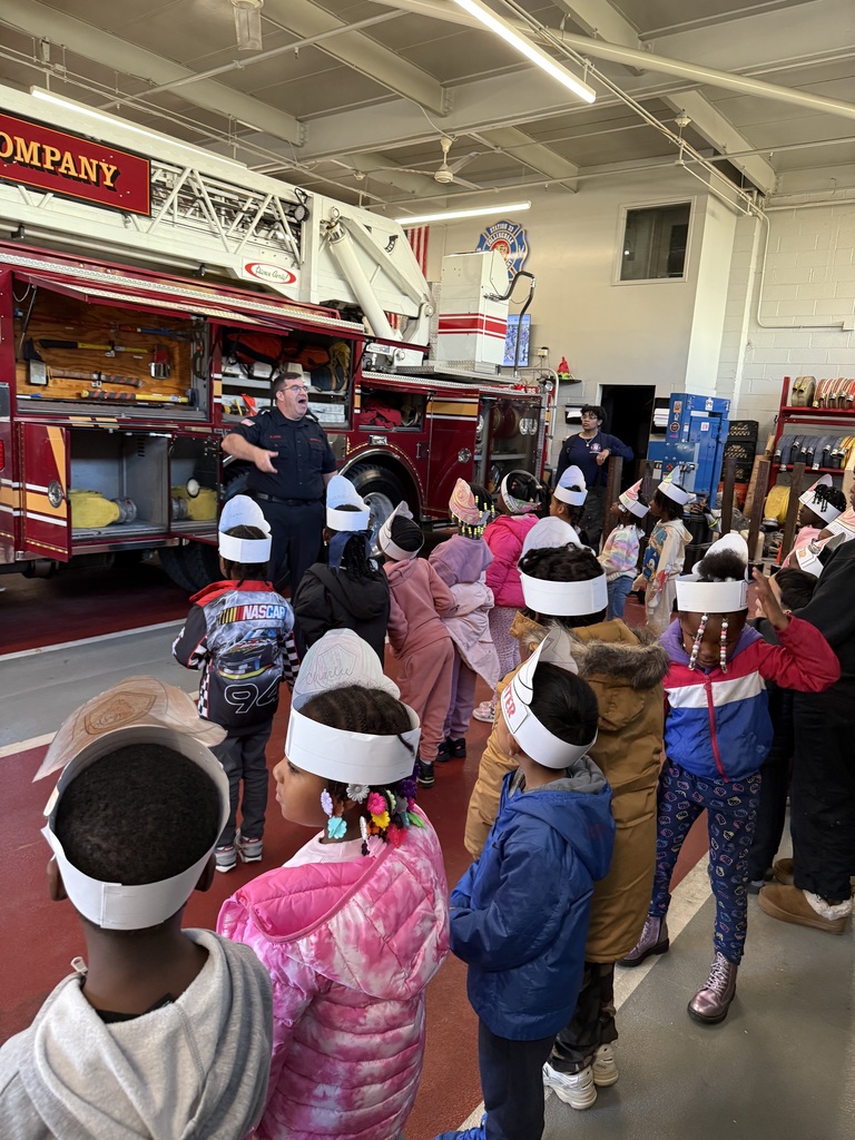 Children wearing paper firefighter hats stand in front of a red fire truck inside the Collingdale Fire Company station, listening to a firefighter giving a presentation.