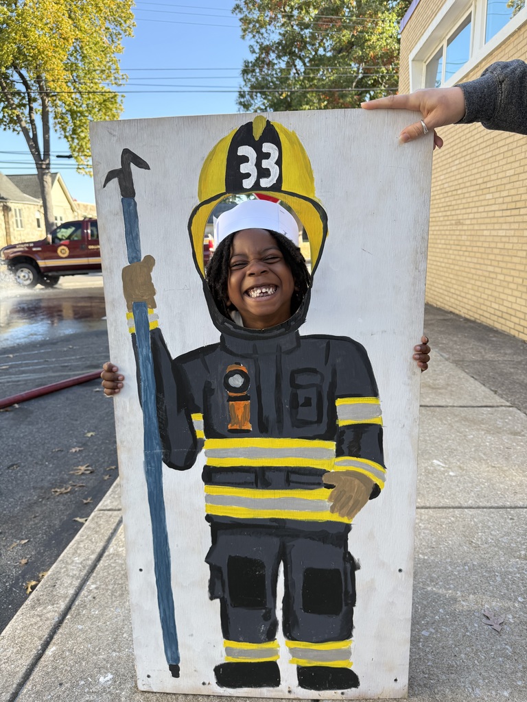 A smiling child stands outdoors, holding a painted board with a firefighter body and helmet labeled “33,” with their face showing through the cutout to appear as the firefighter.
