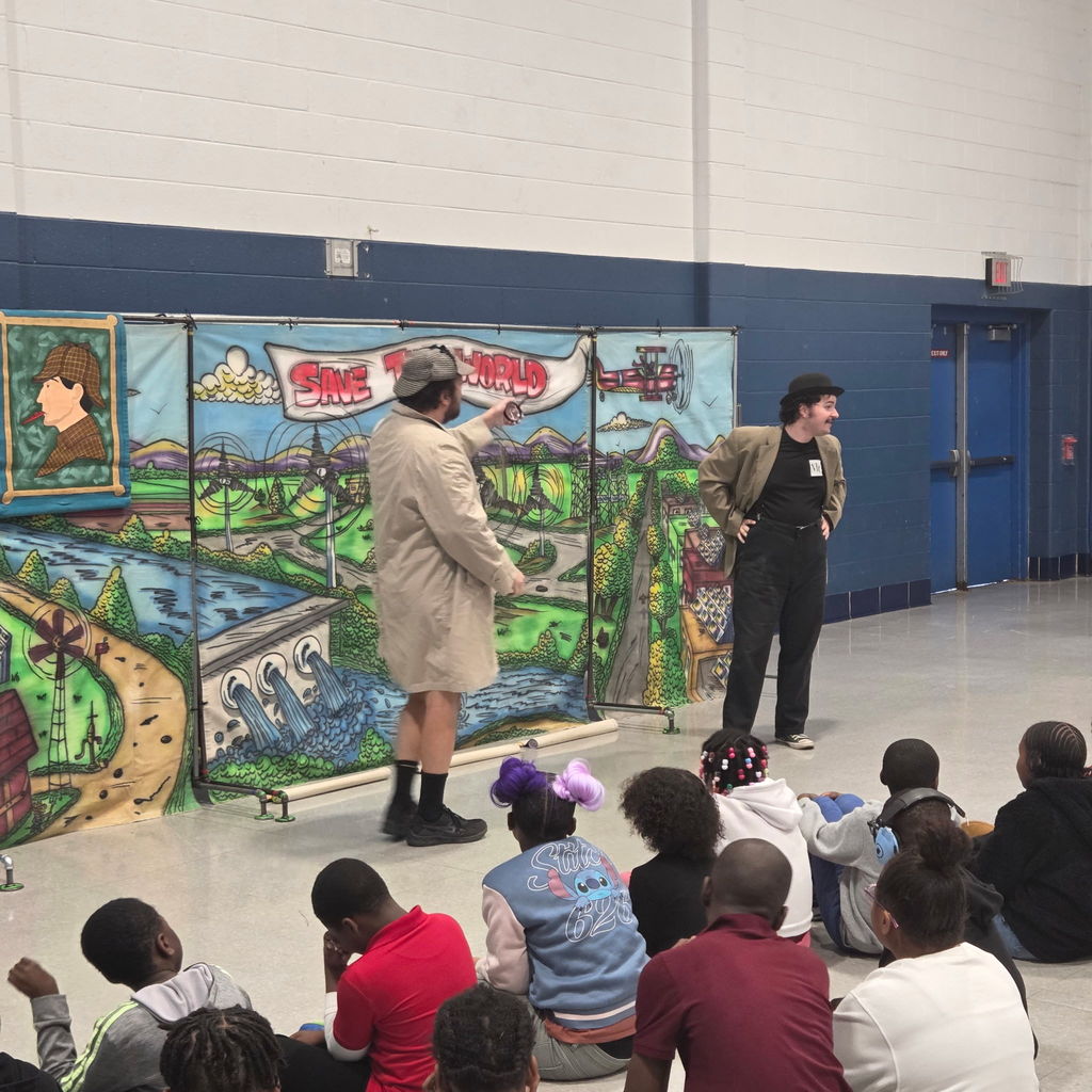 Two performers act out a scene in front of a “Save the World” backdrop, wearing detective-style costumes. One points toward the banner while the other stands with hands on hips. Students sit on the floor in front watching.