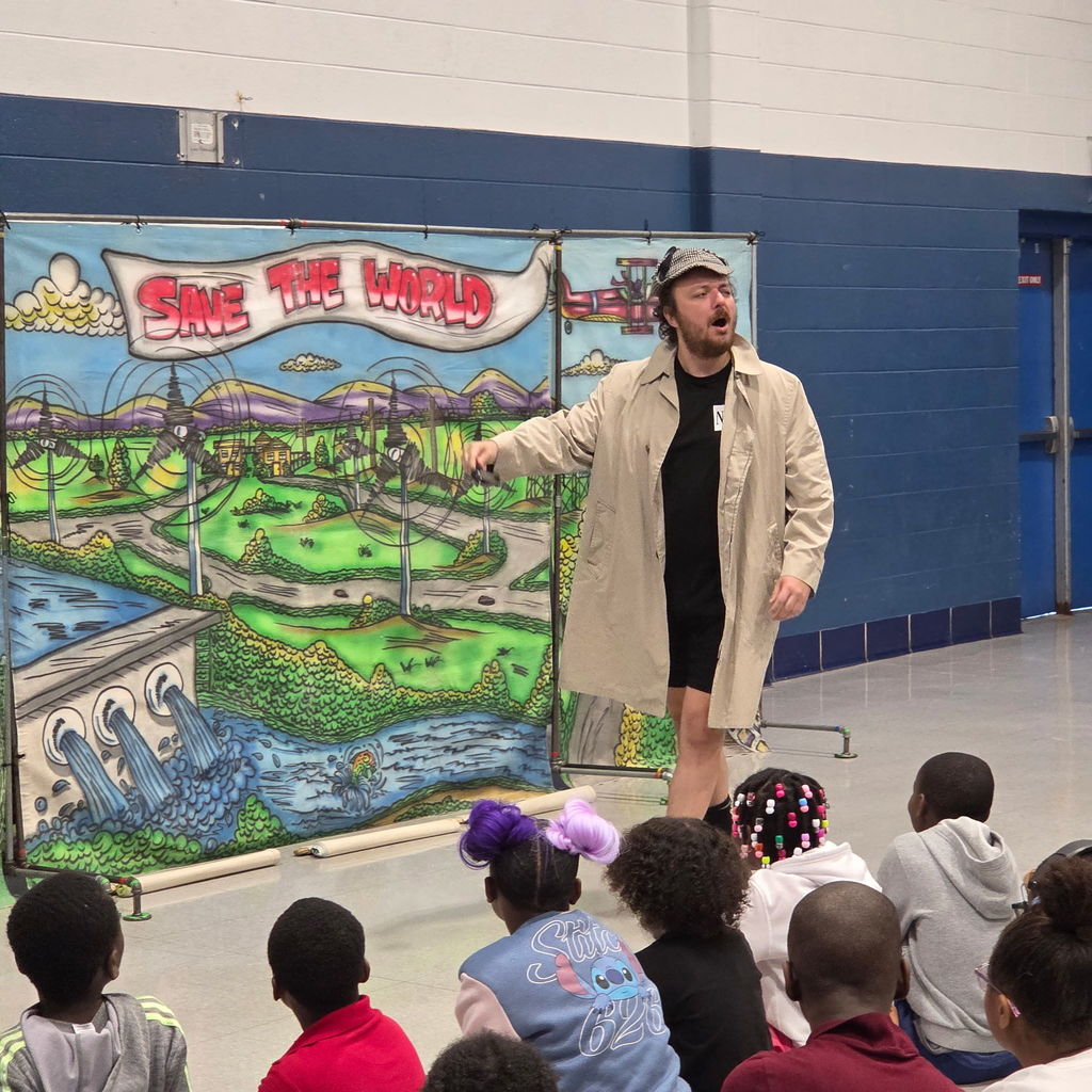 Performer wearing a beige trench coat and detective-style hat gestures dramatically in front of a colorful backdrop reading “Save the World,” featuring illustrations of landscapes, rivers, and buildings. A group of students sits on the floor watching.