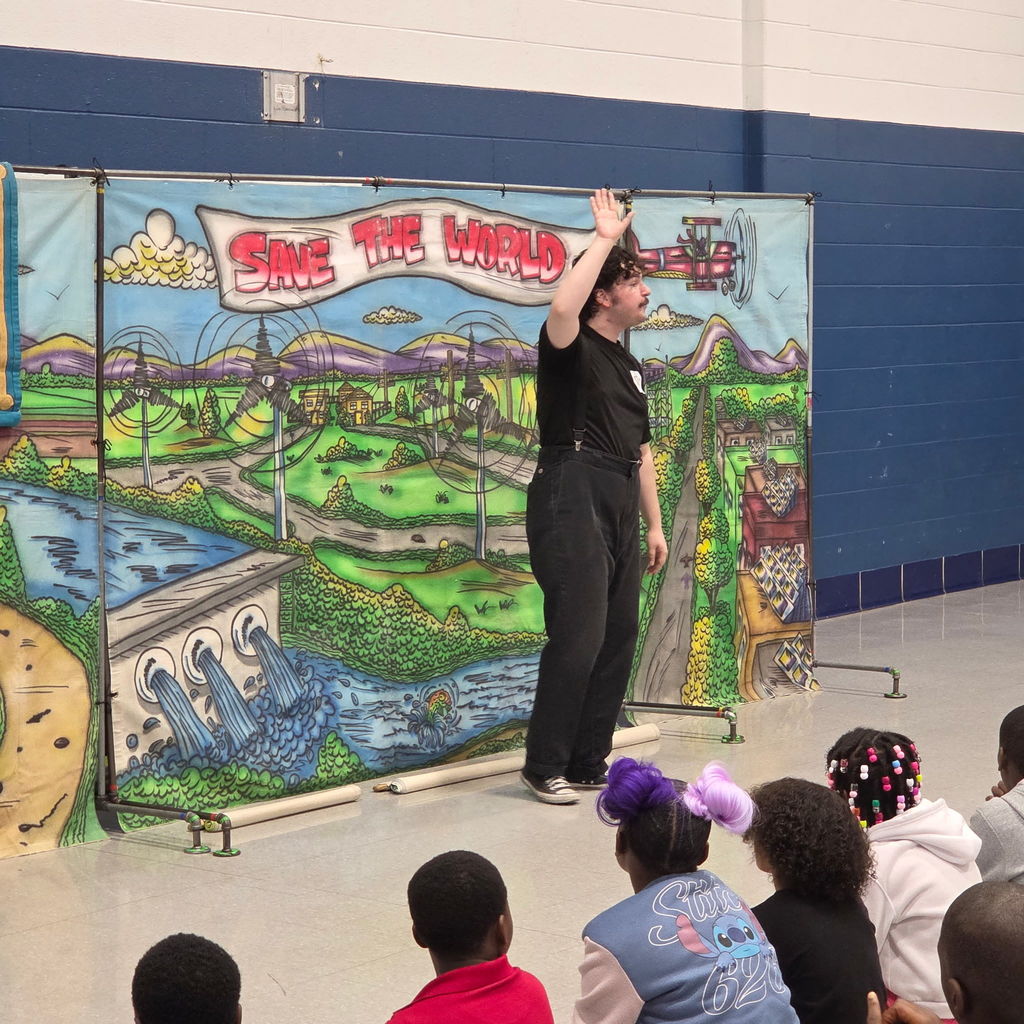 Performer wearing black clothing and suspenders raises a hand while speaking in front of a colorful “Save the World” backdrop. Students sit on the floor facing the stage.