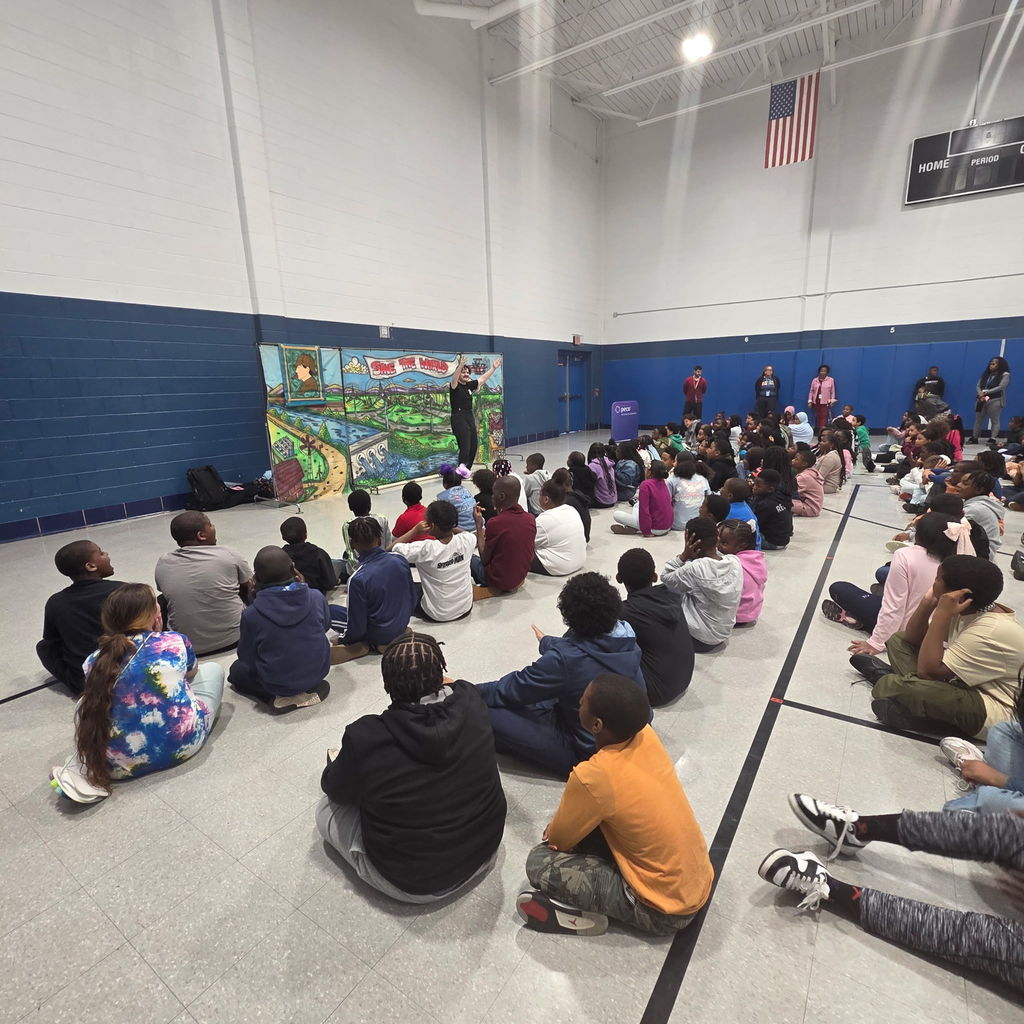 A large group of students sits cross-legged in a school gym, watching a live performance in front of a colorful “Save the World” backdrop.