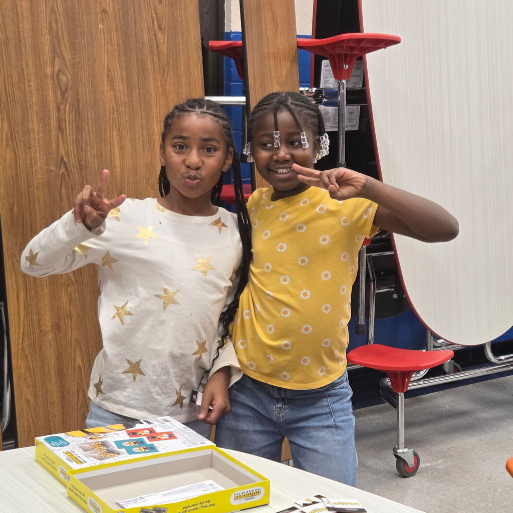 Two children stand side by side, smiling and flashing peace signs. One wears a white shirt with gold stars and the other wears a yellow shirt with small white flowers.