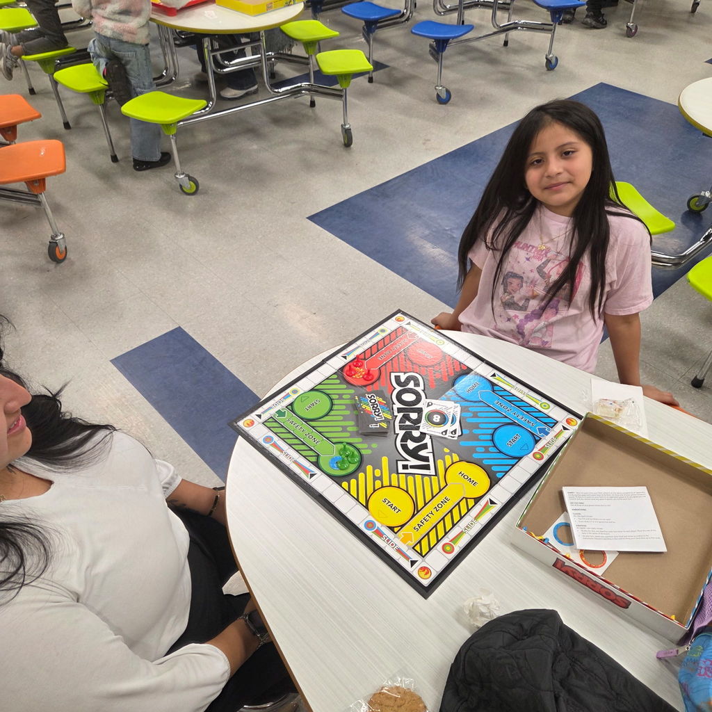 A child and adult sit at a cafeteria table playing the board game Sorry! The child smiles at the camera while the adult looks on. A cookie and game pieces are visible on the table.