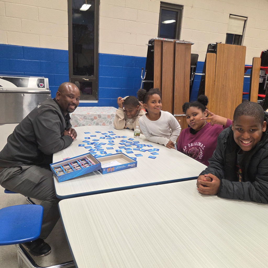 An adult and four children sit around a cafeteria table playing a blue card game. Everyone smiles, and two children make peace signs for the camera.