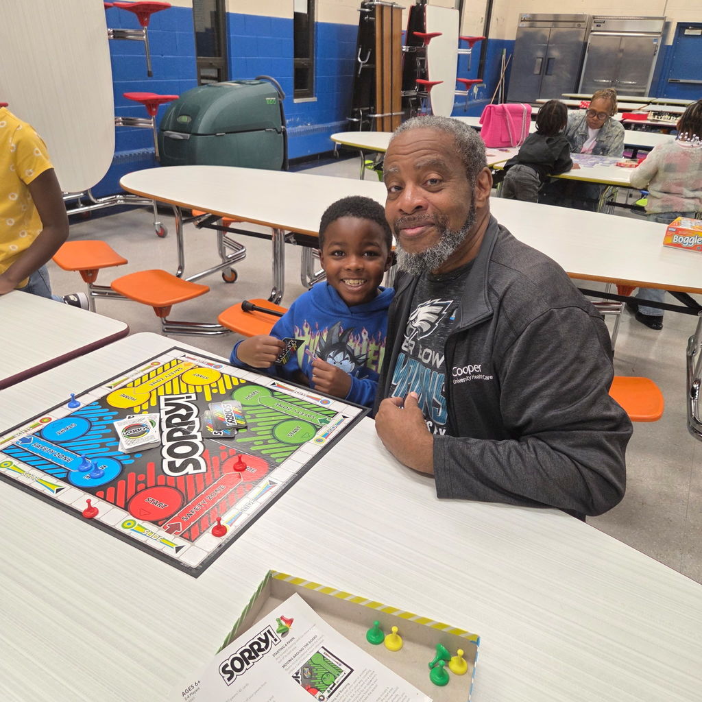 A smiling child and an adult sit at a cafeteria table playing the board game Sorry! together. The child holds a game card, and both look happy as they pose for the photo.