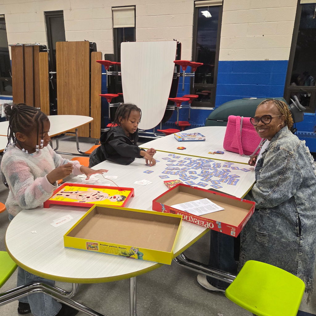 Two children and an adult sit at a cafeteria table playing board games. One child plays Operation while the others play a matching card game. The adult smiles toward the camera.