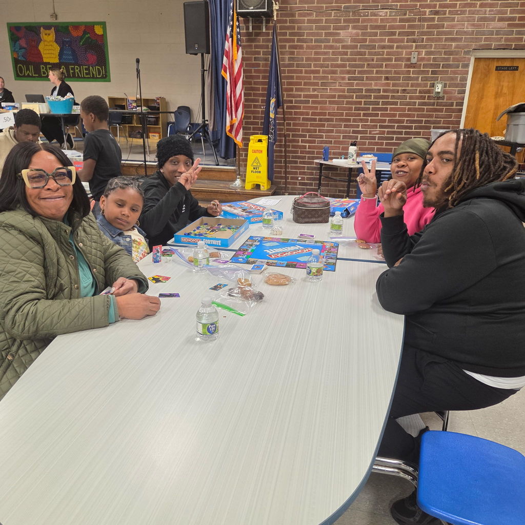 A family sits together at a cafeteria table playing board games, including Monopoly: Fortnite Edition. Some family members smile and make peace signs toward the camera.