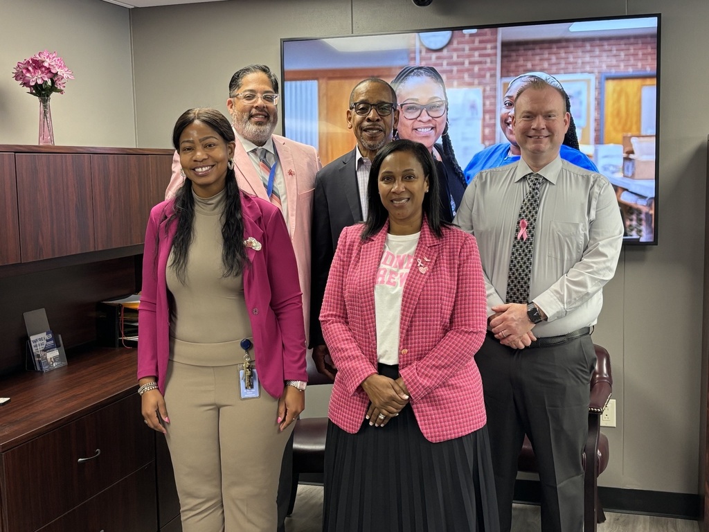 Five staff members stand together in an office, smiling for the photo. They are all wearing pink attire or pink accessories in recognition of Breast Cancer Awareness Month