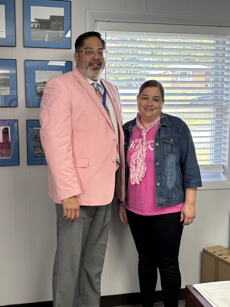Two staff members stand side by side in an office, both wearing pink to support Breast Cancer Awareness Month. The person on the left wears a light pink blazer, while the person on the right wears a pink shirt with a pink scarf and a denim jacket.