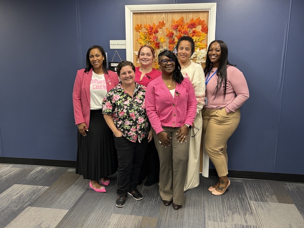Six staff members stand together in front of a blue wall and an autumn-themed door decorated with leaves. They are dressed in pink clothing and accessories, showing support for Breast Cancer Awareness Month.